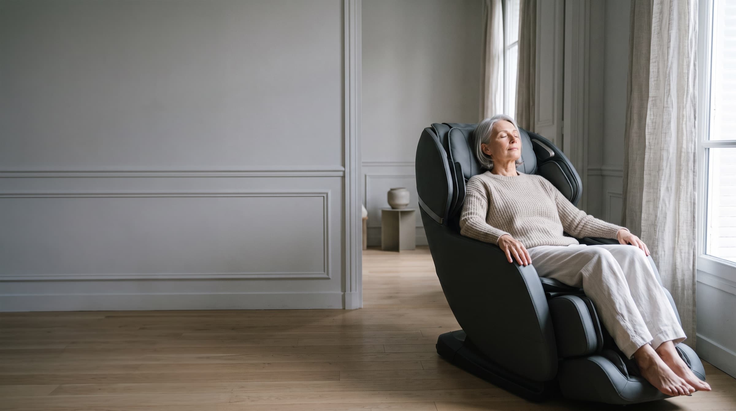 Une femme aux cheveux argentés se détend, les yeux fermés, dans un fauteuil de massage gris graphite, au fond d'un salon clair baigné de lumière